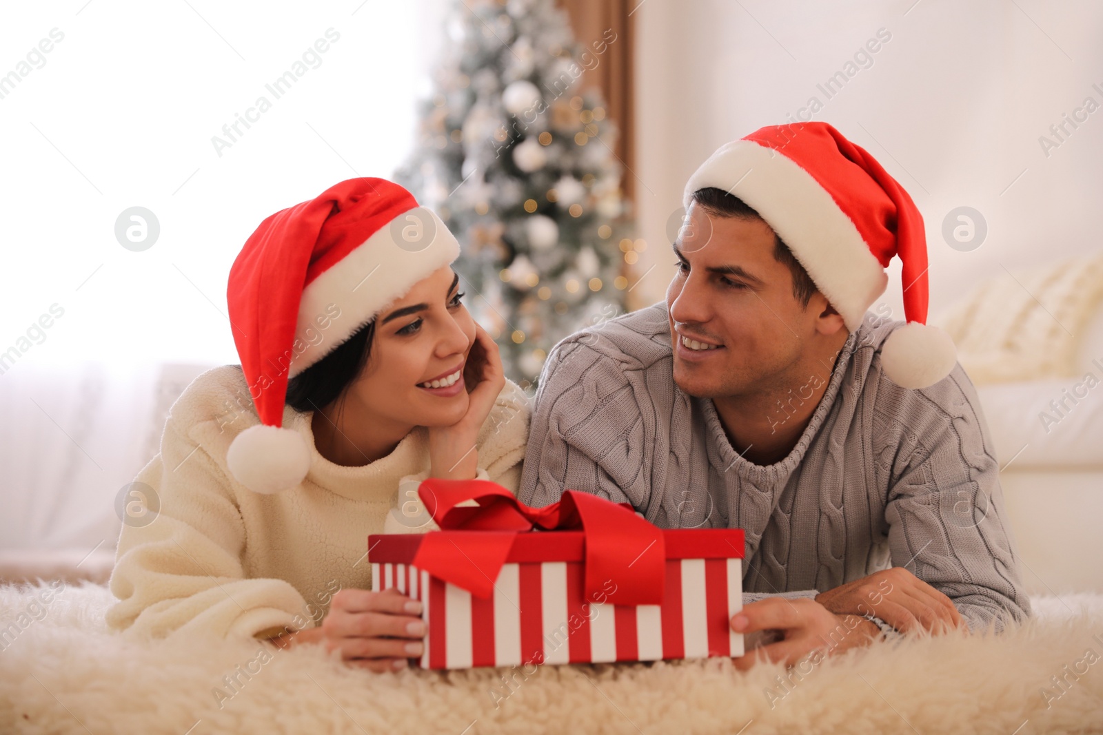 Photo of Couple in Santa hats holding Christmas gift box at home