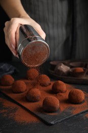Woman covering delicious chocolate truffles with cocoa powder at black table, closeup