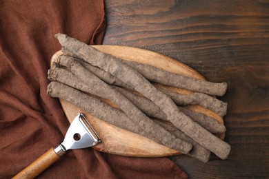 Raw salsify roots and peeler on wooden table, flat lay