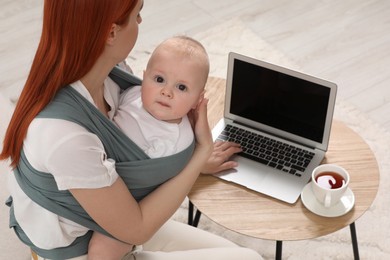 Mother using laptop while holding her child in sling (baby carrier) indoors