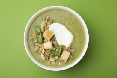 Photo of Delicious broccoli cream soup with croutons, sour cream and pumpkin seeds on green background, top view