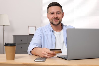 Handsome man with credit card using laptop for online shopping at wooden table in room