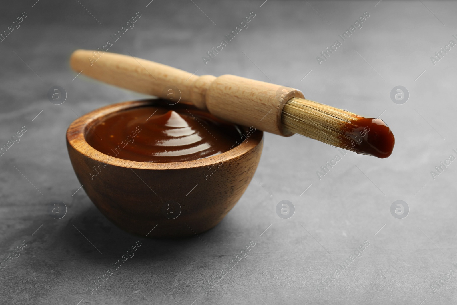 Photo of Tasty barbeque sauce in bowl and brush on grey textured table, closeup