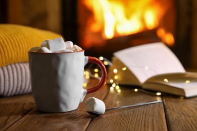 Photo of Mug with hot cocoa, marshmallows, lights and book on wooden table near fireplace