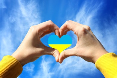 Image of Stop war in Ukraine. Woman showing heart shaped symbol with colors of Ukrainian flag against blue sky outdoors on sunny day, closeup