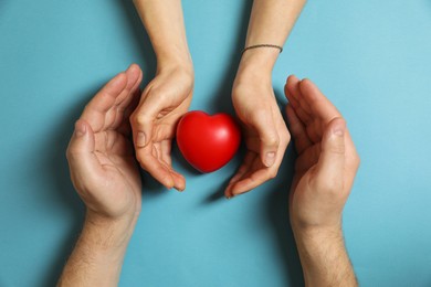 Couple protecting red decorative heart on light blue background, top view