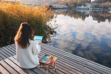Photo of Woman drawing with soft pastels on wooden pier near water, back view
