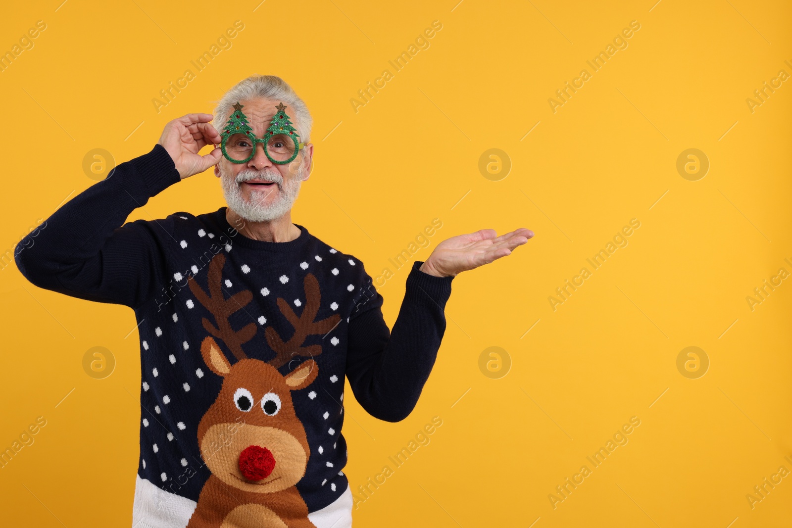 Photo of Senior man in Christmas sweater and funny glasses showing something on orange background. Space for text
