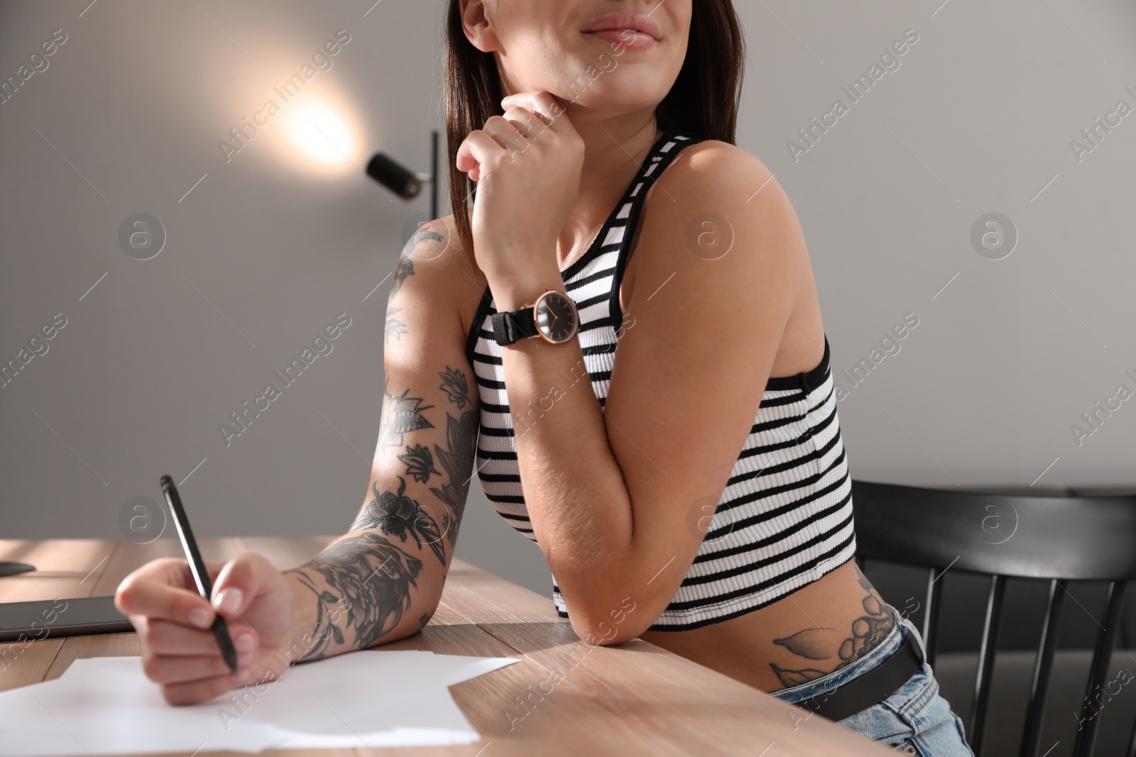 Photo of Beautiful woman with tattoos on body drawing in sketchbook at table indoors, closeup