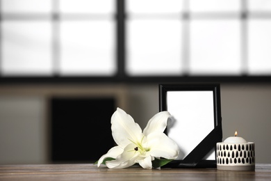Photo of Funeral photo frame with black ribbon, lily and candle on wooden table indoors. Space for design