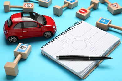 Photo of Many different miniature road signs, notebook with sketch of roundabout and toy car on light blue background. Driving school