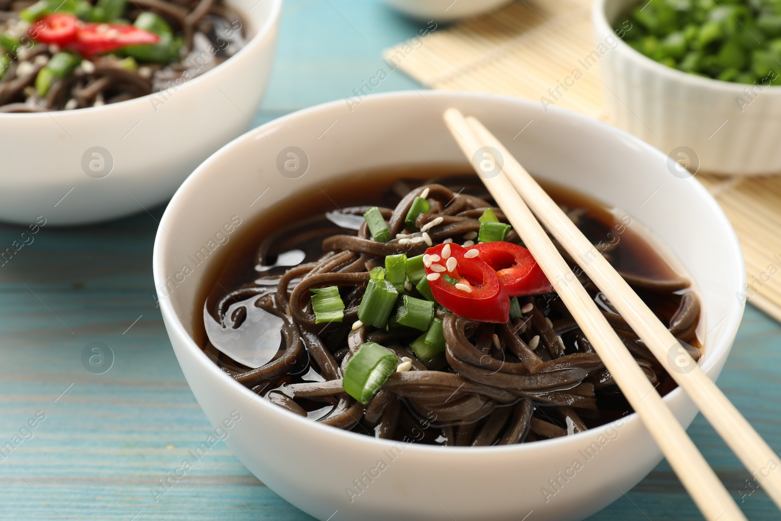 Photo of Tasty soup with buckwheat noodles (soba), chili pepper, green onion in bowl and chopsticks on light blue wooden table, closeup