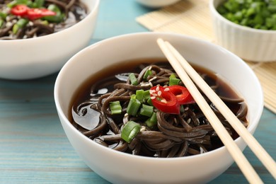 Photo of Tasty soup with buckwheat noodles (soba), chili pepper, green onion in bowl and chopsticks on light blue wooden table, closeup