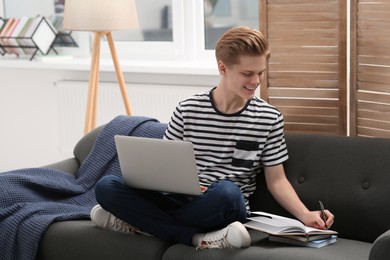 Photo of Online learning. Smiling teenage boy with laptop writing in notebook on sofa at home