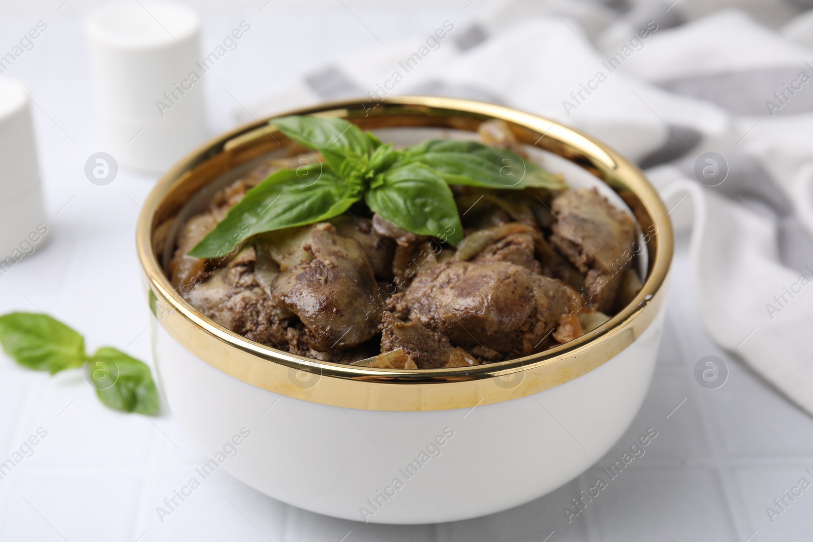 Photo of Delicious fried chicken liver with onion and basil in bowl on white tiled table, closeup