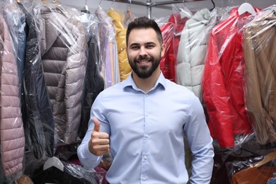 Dry-cleaning service. Happy worker showing thumb up near rack with different clothes indoors