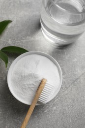Bamboo toothbrush, green leaf, glass of water and bowl of baking soda on grey table, flat lay