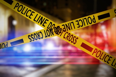 Image of Yellow law enforcement tape isolating crime scene. Blurred view of city street, toned in red and blue police car lights