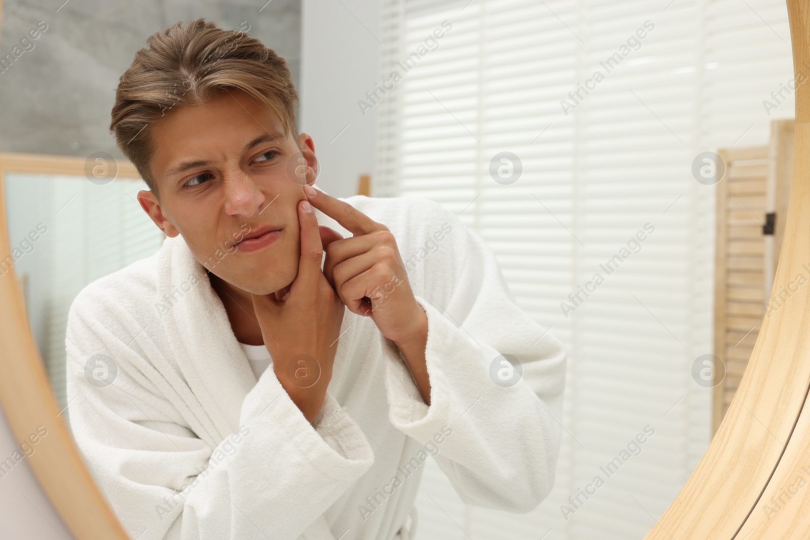 Photo of Upset young man looking at mirror and popping pimple on his face indoors. Acne problem