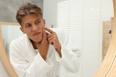 Photo of Upset young man looking at mirror and popping pimple on his face indoors. Acne problem