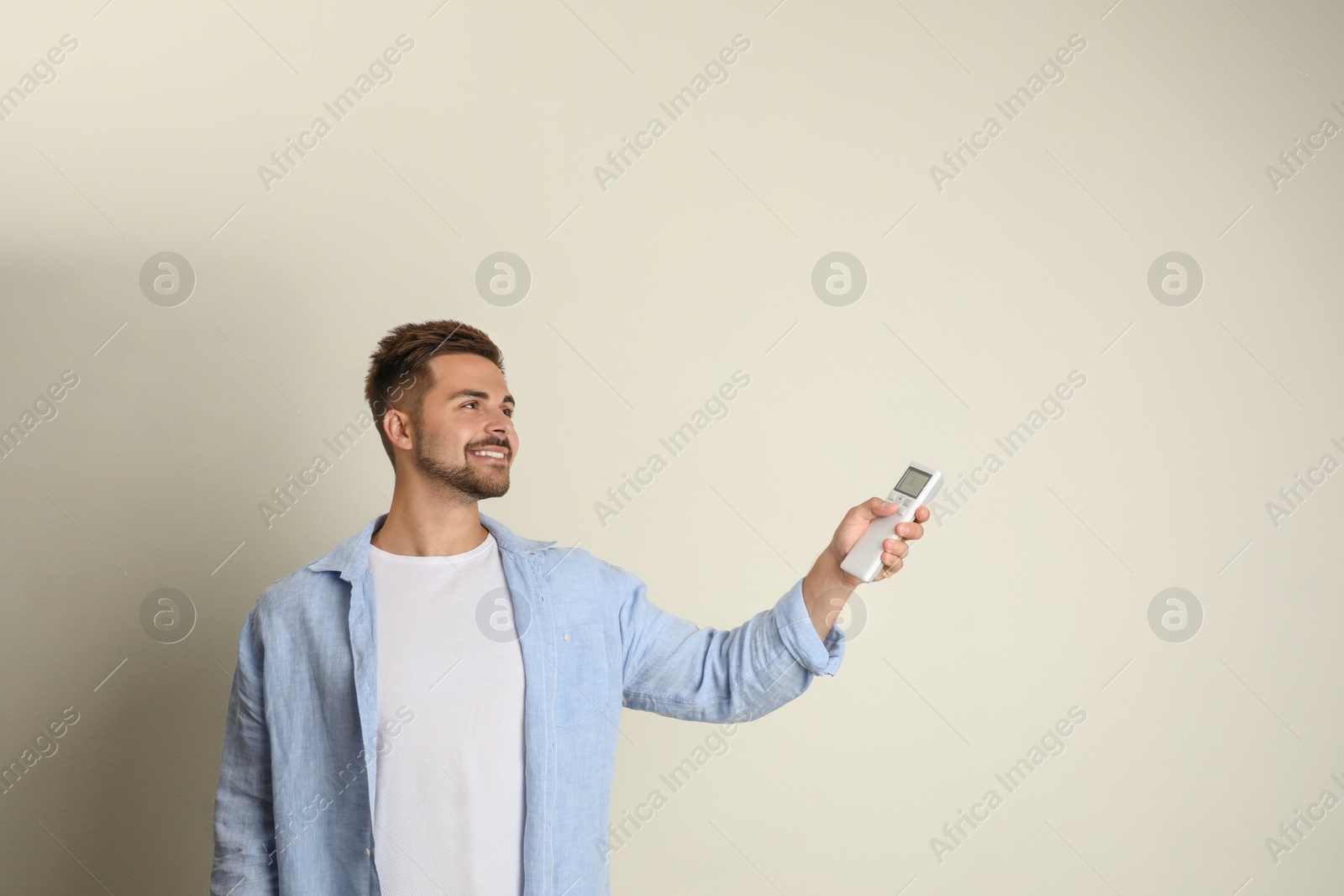 Photo of Happy young man operating air conditioner with remote control on beige background. Space for text