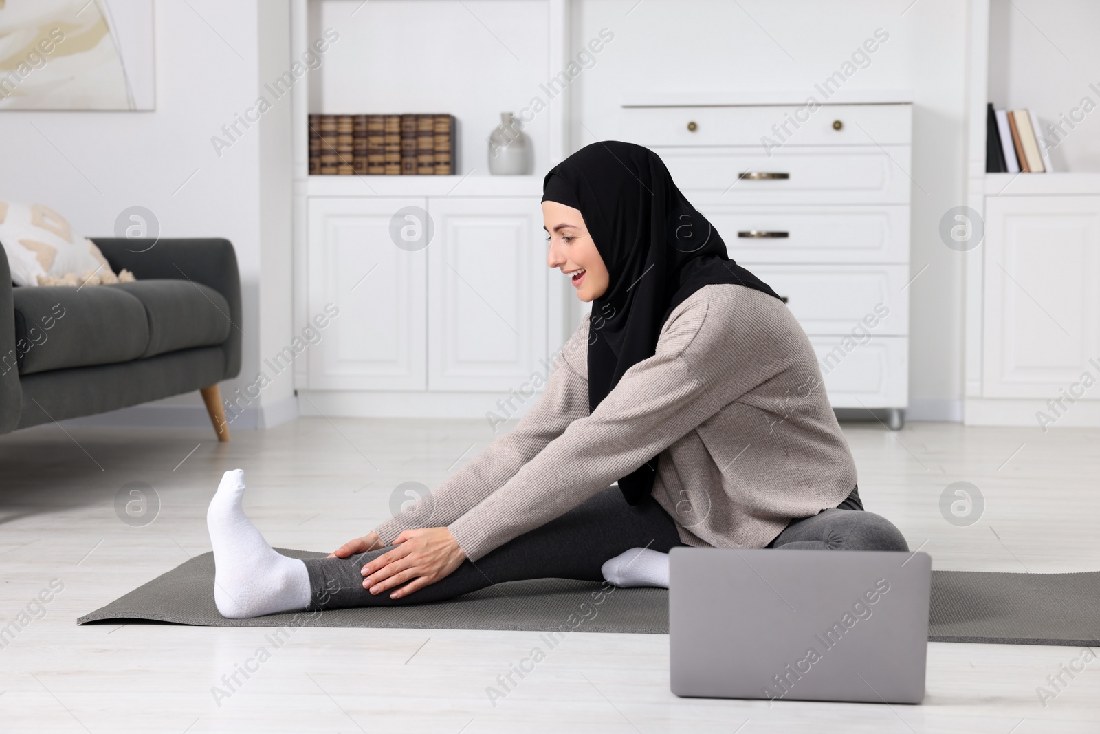 Photo of Muslim woman in hijab stretching near laptop on fitness mat at home