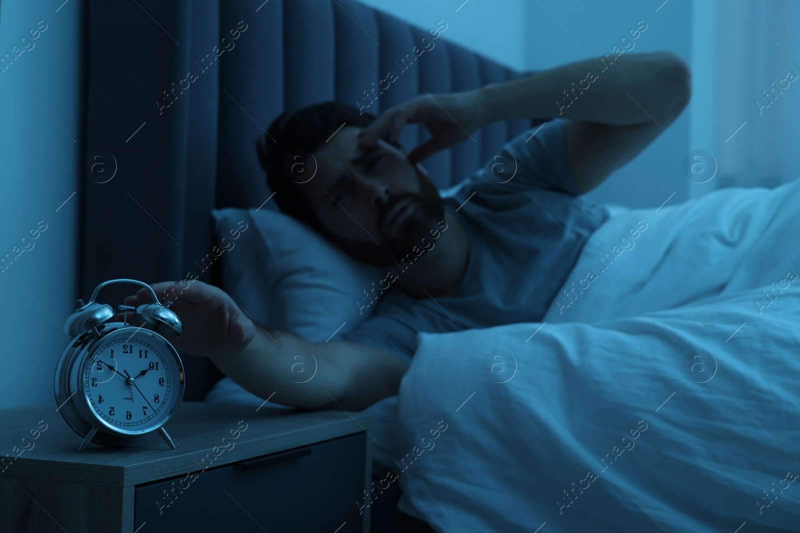 Photo of Man suffering from headache and turning off alarm clock in bed at night, selective focus