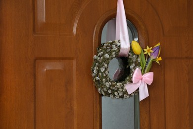 Wreath made of beautiful willow branches, pink bow and colorful tulip flowers on wooden door, space for text