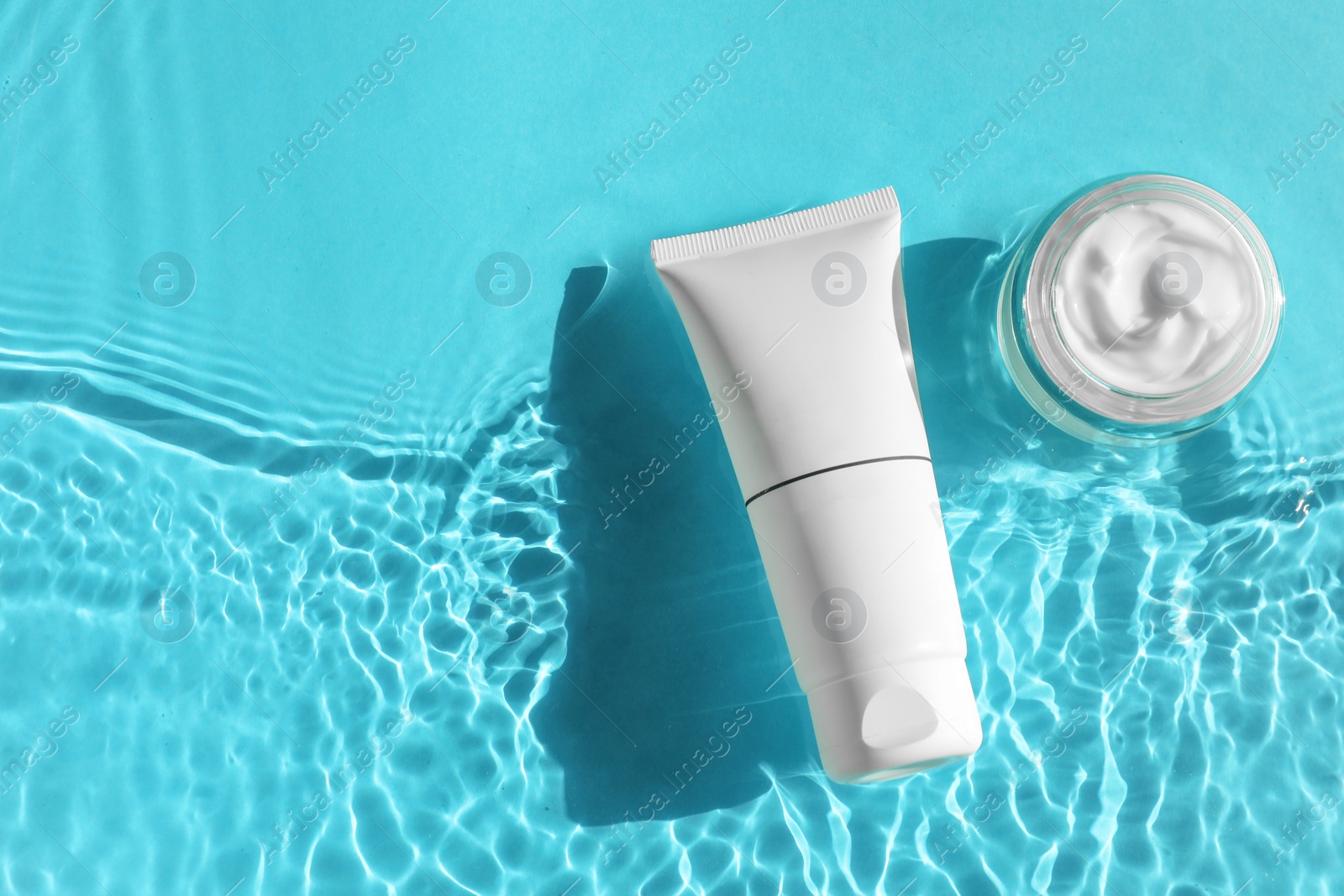 Photo of Tube and jar with moisturizing cream in water on light blue background, top view. Space for text