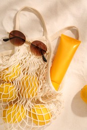 String bag with sunglasses, lemons and sunscreen on sand, flat lay