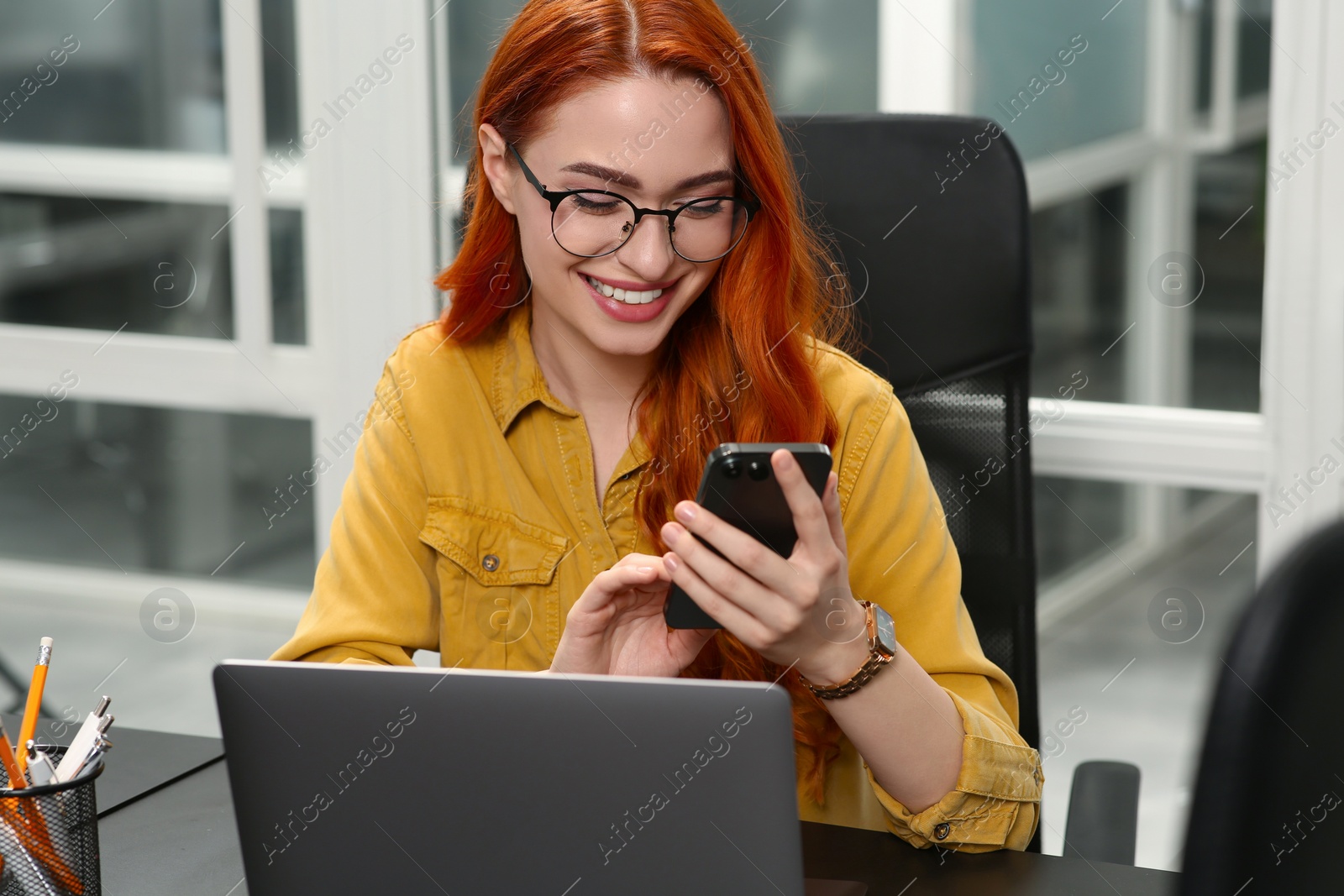 Photo of Happy woman with smartphone working near laptop in office