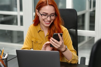 Happy woman with smartphone working near laptop in office
