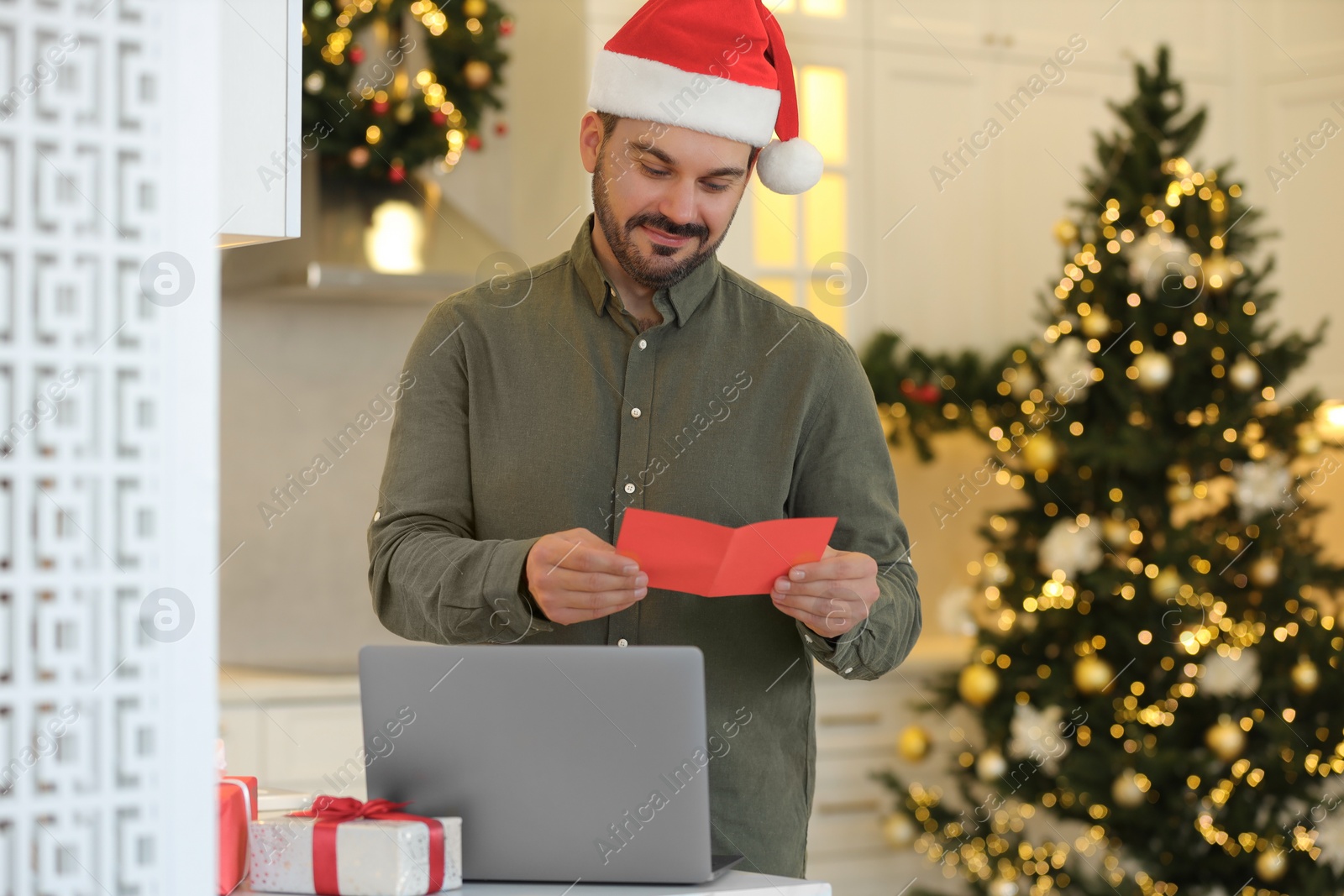 Photo of Celebrating Christmas online with exchanged by mail presents. Man in Santa hat reading greeting card during video call on laptop at home