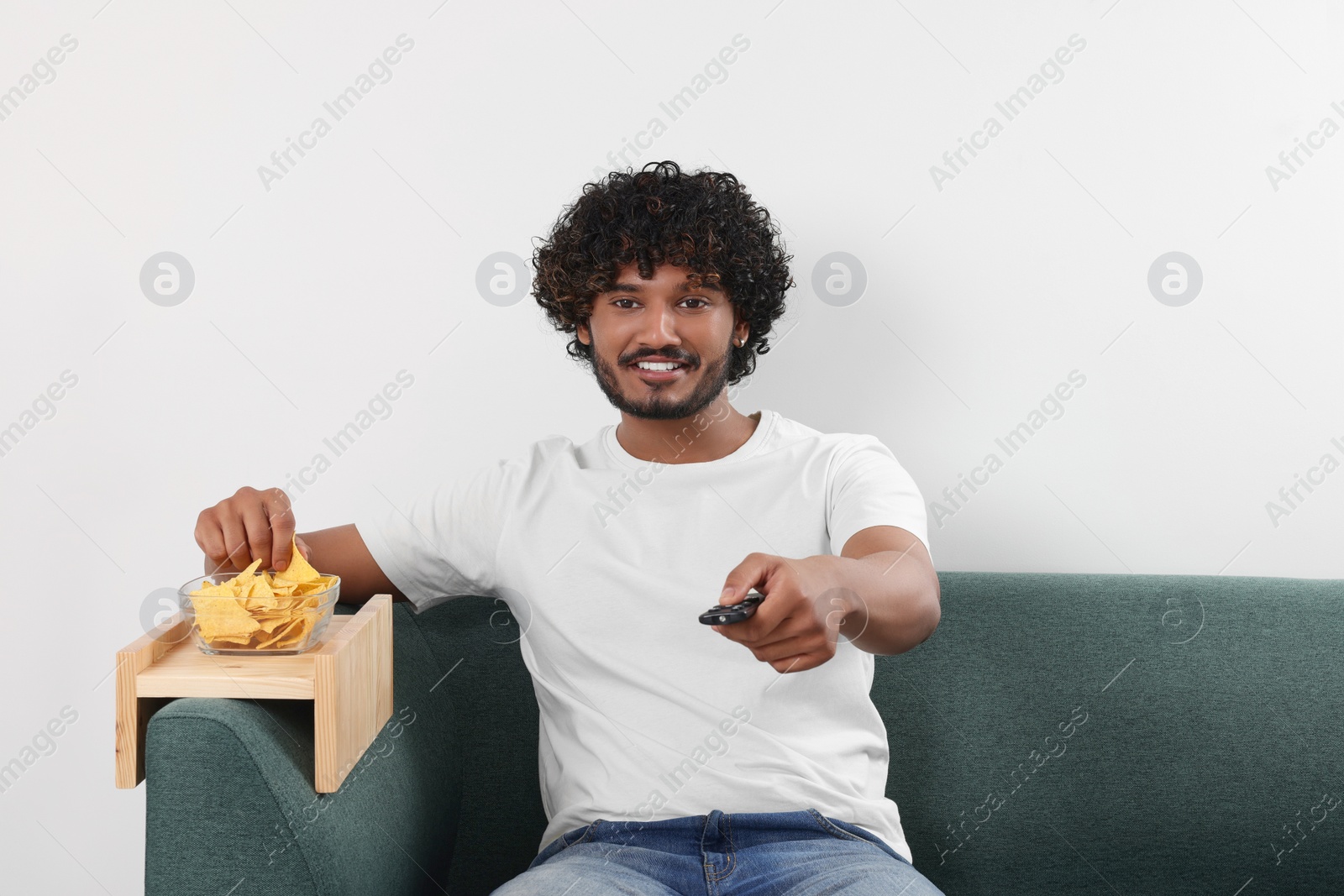 Photo of Happy man switching TV channels while eating nacho chips on sofa with wooden armrest table at home