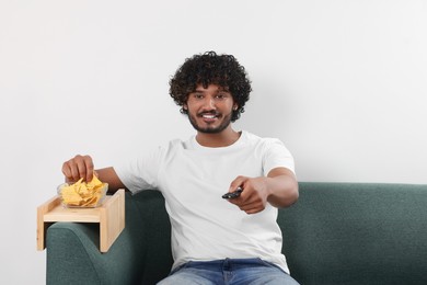 Happy man switching TV channels while eating nacho chips on sofa with wooden armrest table at home