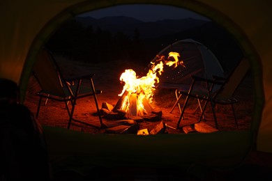 Photo of Beautiful bonfire and folding chairs outdoors in evening, view from camping tent