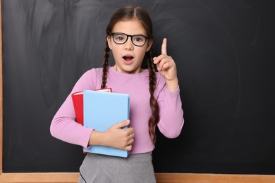 Photo of Surprised schoolgirl in glasses holding books near chalkboard