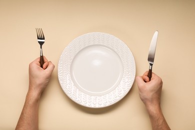 Photo of Man holding fork and knife near empty plate at beige table, top view