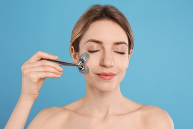 Young woman using metal face roller on light blue background