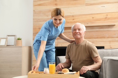 Photo of Nurse serving breakfast to elderly man indoors. Assisting senior people