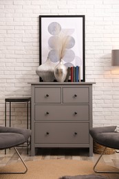 Stylish room interior with grey chest of drawers and chairs near white brick wall