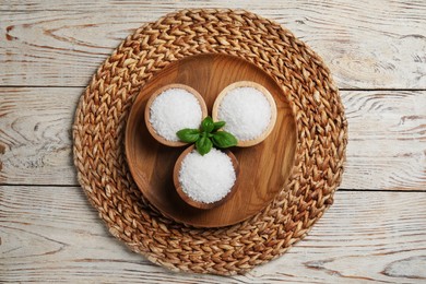 Photo of Bowls of natural sea salt and wicker mat on white wooden table, flat lay