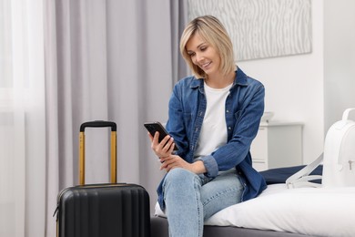 Smiling guest with smartphone relaxing on bed in stylish hotel room
