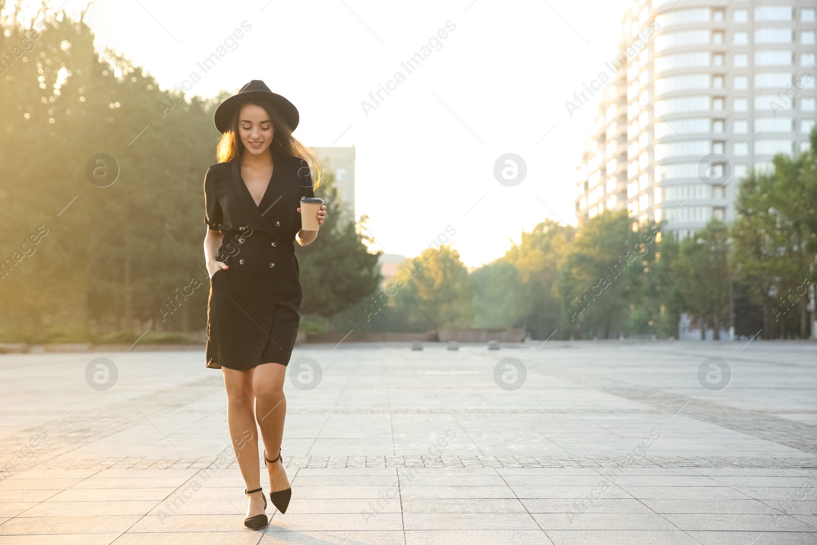 Photo of Beautiful young woman in stylish black dress and hat with cup of coffee on city street