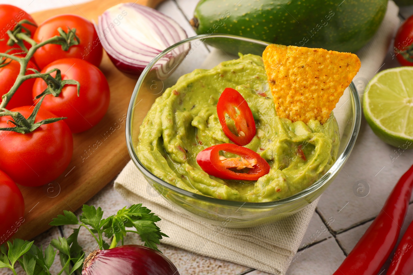 Photo of Bowl of delicious guacamole, nachos chip and ingredients on white tiled table, closeup