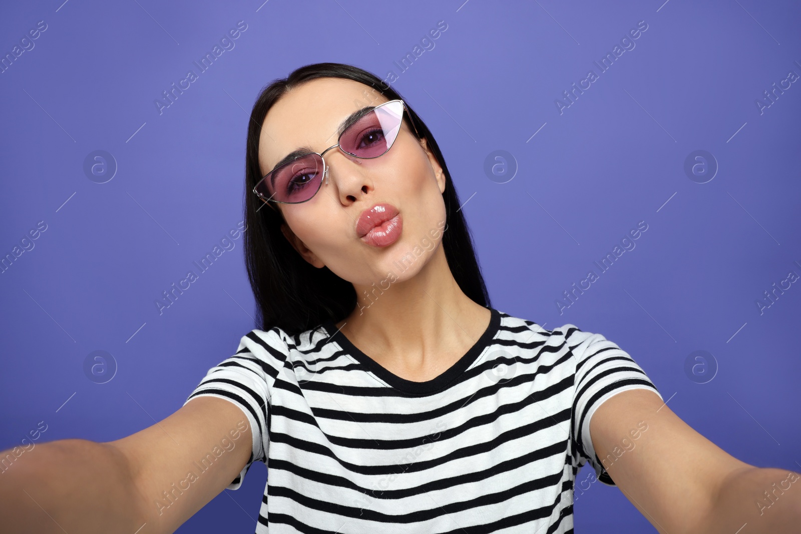 Photo of Beautiful young woman in stylish sunglasses taking selfie while blowing kiss on purple background