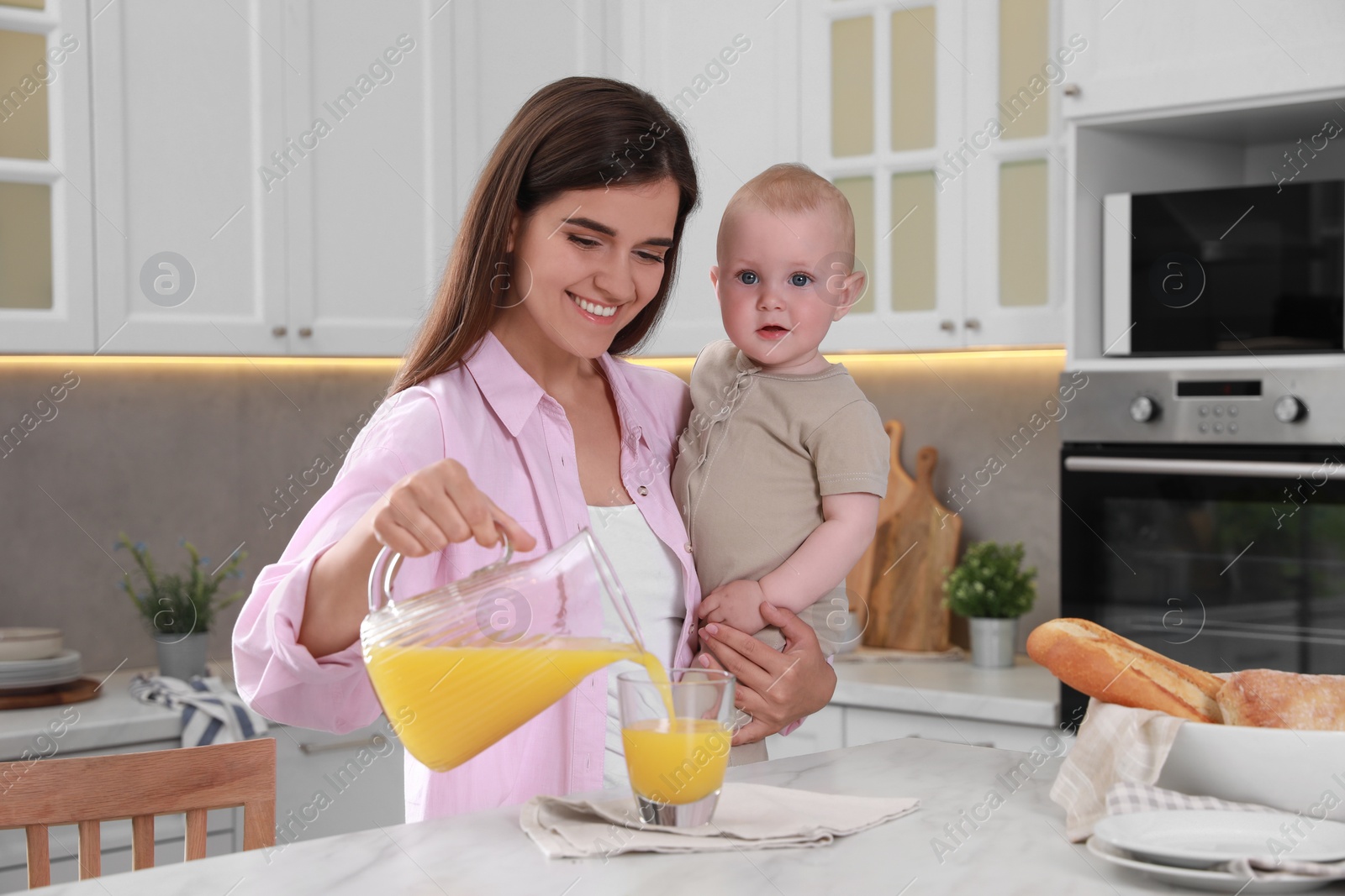 Photo of Happy young woman holding her cute little baby while pouring juice into glass in kitchen
