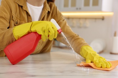 Photo of Woman with spray bottle and microfiber cloth cleaning white marble table in kitchen, closeup