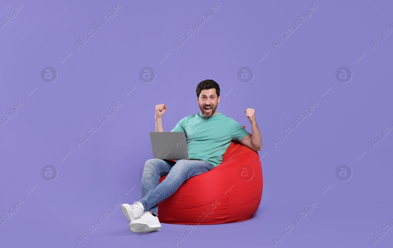 Photo of Emotional man with laptop sitting on beanbag chair against purple background