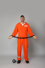 Prisoner in orange jumpsuit with chained hands and metal ball on grey background